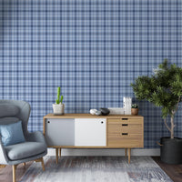 A contemporary living room with blue plaid wallpaper creating a textured backdrop. The space features a gray upholstered armchair, a mid-century modern wooden sideboard with decorative objects and books, and a large potted indoor tree on dark wood flooring.
