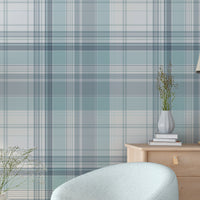 A bright room features a light blue and white plaid wallpaper pattern.
The wallpaper colors include shades of soft aqua, dusty blue, and white lines.
A rounded light blue armchair sits on the dark wood floor.
A small, round wooden side table holds a white vase with flowers.
A large green plant in a white pot stands on the left side.
A light wood dresser with a small desk lamp sits on the right.