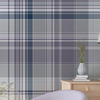A stylish living room featuring a large plaid-patterned wallpaper in soft lilac, gray, and navy tones. The space includes a pale lavender accent chair, a round wooden side table with a small vase of flowers, a wooden cabinet with decor, and a tall green potted plant on dark wood flooring.