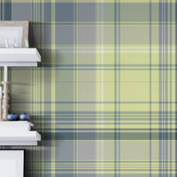 A modern interior featuring green and blue plaid wallpaper with a soft tartan pattern. The room includes floating white shelves with decorative objects, a light wood chair, and a small round table with a black adjustable desk lamp on a light wood floor.