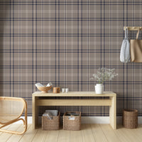 A cozy entryway setup is styled against a taupe, navy, and white plaid wallpaper background.
A long, light-colored wooden console table holds a bowl of books, a small round box, candles, and a white vase with delicate flowers.
Tucked neatly beneath the table are two woven storage baskets filled with magazines and reading materials.