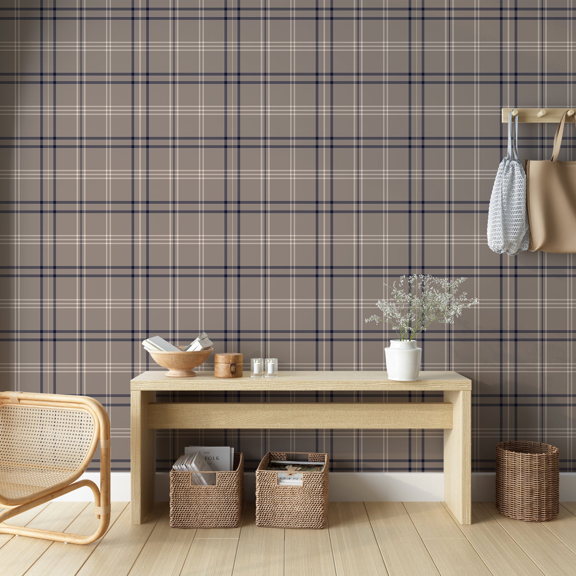 A cozy entryway setup is styled against a taupe, navy, and white plaid wallpaper background.
A long, light-colored wooden console table holds a bowl of books, a small round box, candles, and a white vase with delicate flowers.
Tucked neatly beneath the table are two woven storage baskets filled with magazines and reading materials.