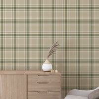 A modern living space featuring a muted wheat-beige, sage green, and white plaid wallpaper accent wall. In the center, a light oak sideboard cabinet with wooden legs holds a small wooden tripod lamp, a geometric candle, and a round white ceramic vase with a single sprig of dried pampas grass. To the left, a woven basket sits on the light wood floor, filled with tall, fluffy pampas grass stalks and a draped blanket.