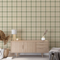 A modern living space featuring a muted wheat-beige, sage green, and white plaid wallpaper accent wall. In the center, a light oak sideboard cabinet with wooden legs holds a small wooden tripod lamp, a geometric candle, and a round white ceramic vase with a single sprig of dried pampas grass. To the left, a woven basket sits on the light wood floor, filled with tall, fluffy pampas grass stalks and a draped blanket.