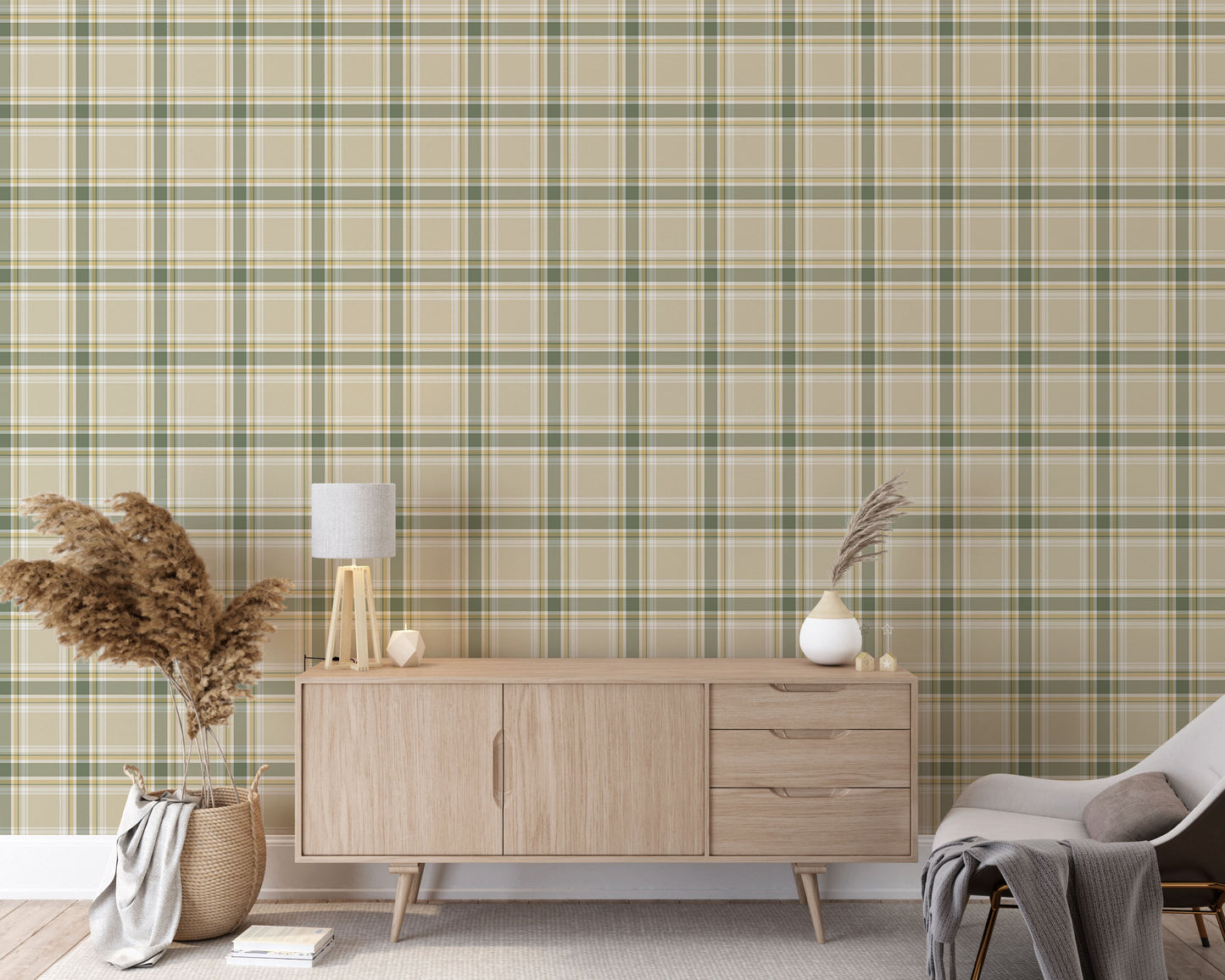 A modern living space featuring a muted wheat-beige, sage green, and white plaid wallpaper accent wall. In the center, a light oak sideboard cabinet with wooden legs holds a small wooden tripod lamp, a geometric candle, and a round white ceramic vase with a single sprig of dried pampas grass. To the left, a woven basket sits on the light wood floor, filled with tall, fluffy pampas grass stalks and a draped blanket.