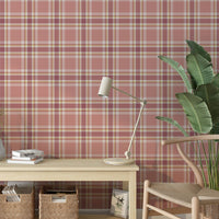 A brightly lit interior space featuring a blushed brick-red and cream plaid wallpaper accent wall. In the center sits a low, light wood console table holding a small white fluted vase with sprigs, a short stack of books, and a sleek cream desk lamp. Underneath the table, two woven wicker baskets store books and magazines. To the left, a woven basket holds tall, decorative dried fan palm leaves, while a modern light wood wishbone chair rests on the right next to a large potted banana leaf plant.