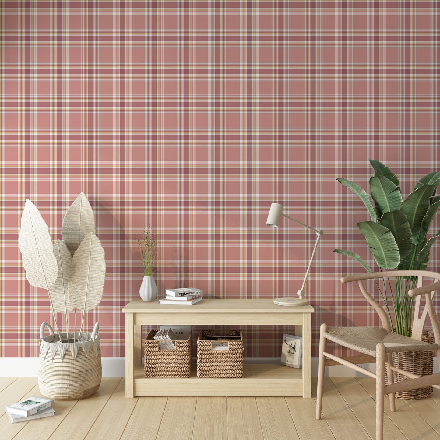 A brightly lit interior space featuring a blushed brick-red and cream plaid wallpaper accent wall. In the center sits a low, light wood console table holding a small white fluted vase with sprigs, a short stack of books, and a sleek cream desk lamp. Underneath the table, two woven wicker baskets store books and magazines. To the left, a woven basket holds tall, decorative dried fan palm leaves, while a modern light wood wishbone chair rests on the right next to a large potted banana leaf plant.