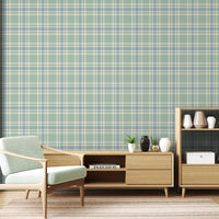 A light and airy living room featuring a misty sage green, soft blue, and pale yellow plaid wallpaper accent wall. In the foreground, a mid-century modern armchair with a pale mint-green cushion sits on a striped area rug. Behind the chair, a long light oak media console with open shelving displays books and potted succulents. To the right stands a matching small light oak bookshelf styled with white, beige, and black ceramic vases and a decorative white box.