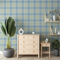 A bright bedroom or living area featuring a light blue, sage green, and white plaid wallpaper accent wall. Centered against the wall is a light birch chest of drawers with simple leather tab pulls, holding a circular white ceramic vase and a book. To the left stands a large potted bird of paradise plant in a low, round grey concrete planter. To the right, a matching two-tiered wooden shelving unit with leather straps hangs on the wall over a small nightstand holding lit candles and an abstract sculpture.