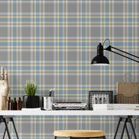 A closeup of a modern desk workspace featuring a soft graphite grey plaid wallpaper with light blue and pale yellow accent lines. In the foreground, a white tabletop rests on black trestle legs, paired with a wooden stool. The desk is styled with a prominent black metal architect-style desk lamp on the right, stacks of design books and magazines, wire storage baskets, a small potted plant, a silver pencil cup, and an articulated wooden mannequin hand decoration on the far left.