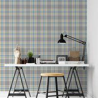 A closeup of a modern desk workspace featuring a soft graphite grey plaid wallpaper with light blue and pale yellow accent lines. In the foreground, a white tabletop rests on black trestle legs, paired with a wooden stool. The desk is styled with a prominent black metal architect-style desk lamp on the right, stacks of design books and magazines, wire storage baskets, a small potted plant, a silver pencil cup, and an articulated wooden mannequin hand decoration on the far left.