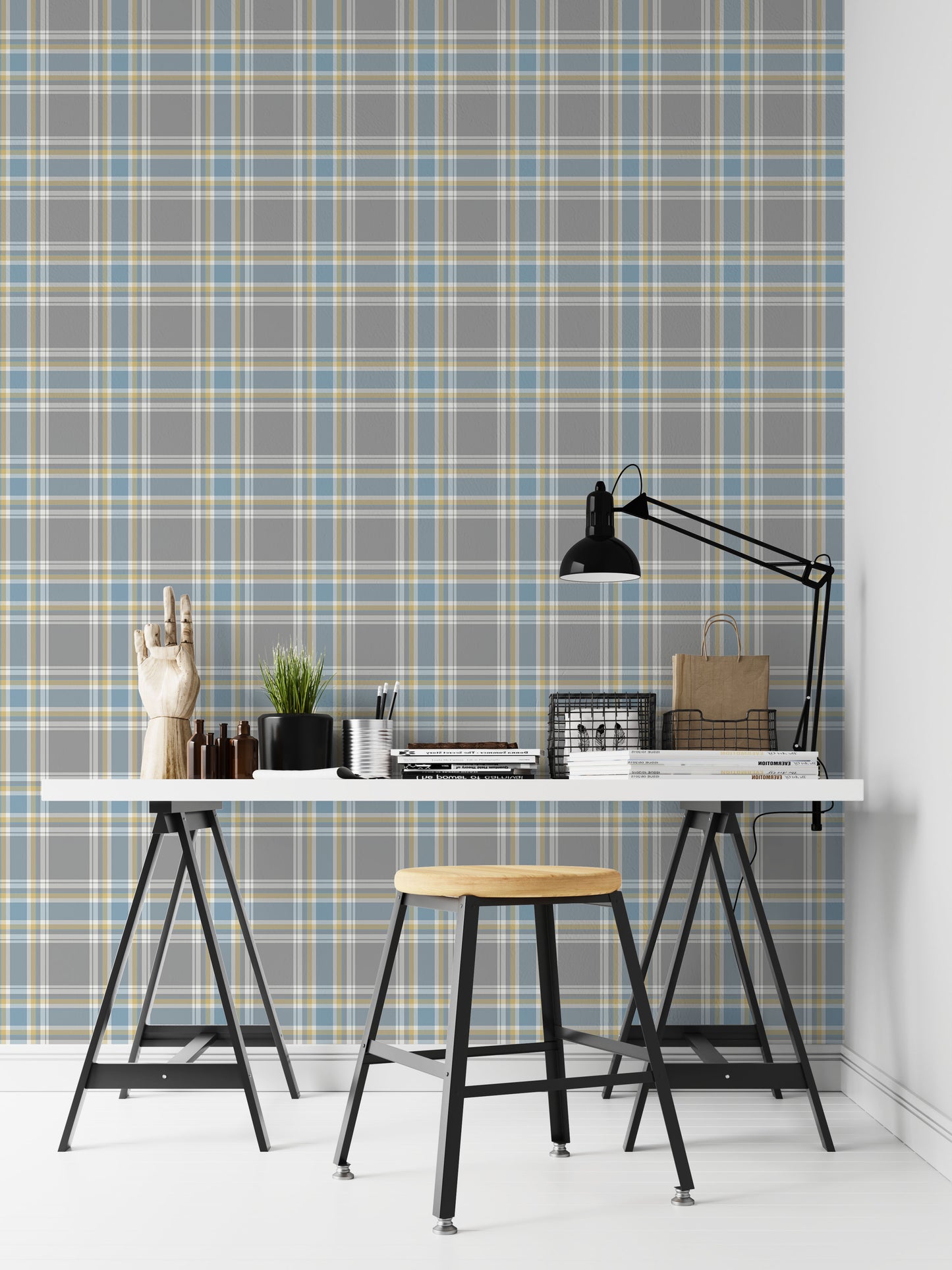A closeup of a modern desk workspace featuring a soft graphite grey plaid wallpaper with light blue and pale yellow accent lines. In the foreground, a white tabletop rests on black trestle legs, paired with a wooden stool. The desk is styled with a prominent black metal architect-style desk lamp on the right, stacks of design books and magazines, wire storage baskets, a small potted plant, a silver pencil cup, and an articulated wooden mannequin hand decoration on the far left.