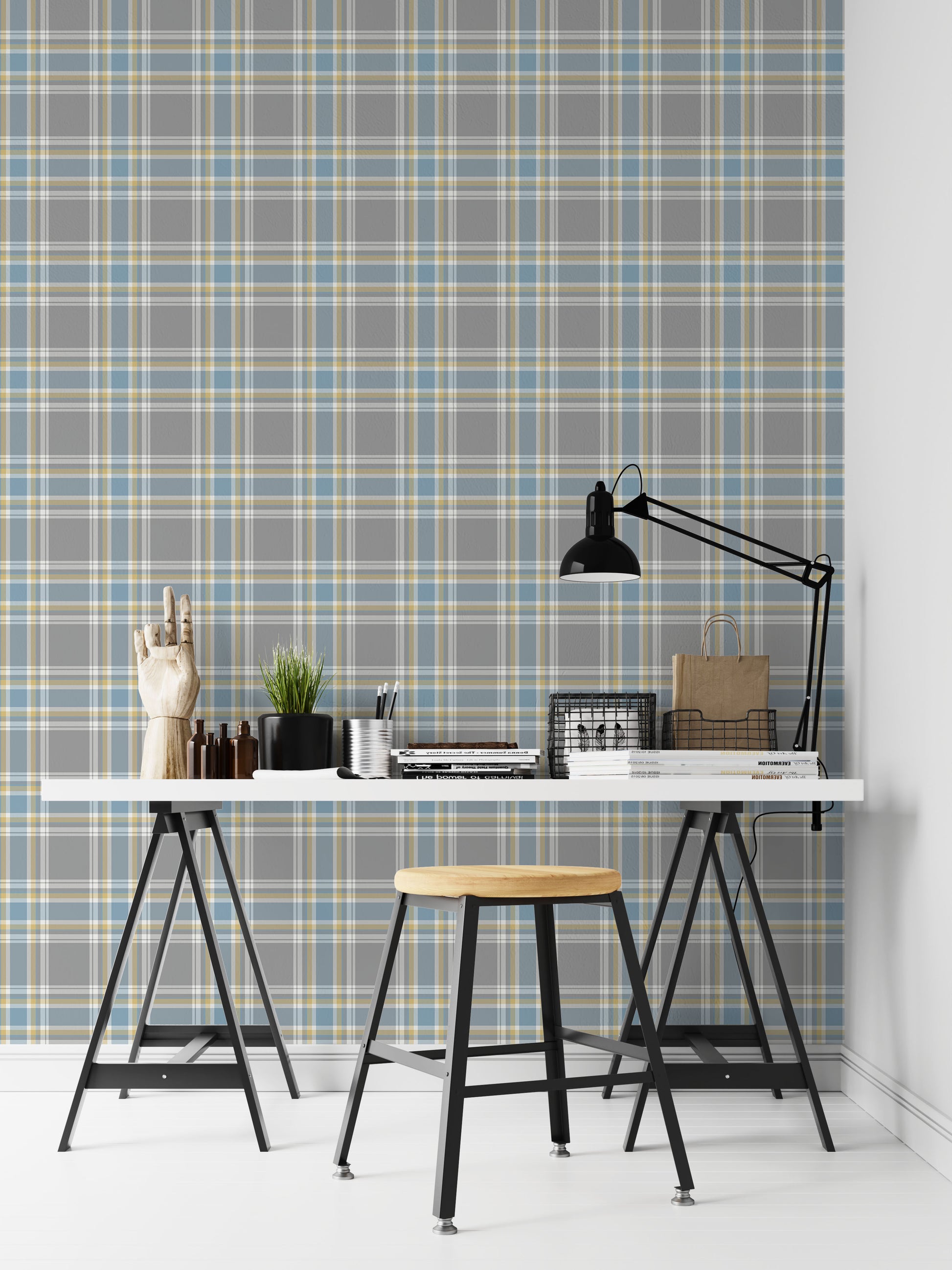 A closeup of a modern desk workspace featuring a soft graphite grey plaid wallpaper with light blue and pale yellow accent lines. In the foreground, a white tabletop rests on black trestle legs, paired with a wooden stool. The desk is styled with a prominent black metal architect-style desk lamp on the right, stacks of design books and magazines, wire storage baskets, a small potted plant, a silver pencil cup, and an articulated wooden mannequin hand decoration on the far left.