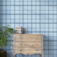 A serene interior space features a classic plaid wallpaper in light steel blue and cream tones.
The textured design displays a detailed grid pattern with intersecting blue, grey, and white lines.
In front, a rustic wooden chest of drawers stands against the wall holding a stack of books.
The charming interior setting is completed by a leafy green potted plant placed on the left side.