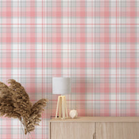 A modern living space featuring a soft cherry blossom pink, white, and grey plaid wallpaper accent wall. In the center, a light oak sideboard cabinet with wooden legs holds a small wooden tripod lamp, a geometric candle, and a round white ceramic vase with a single sprig of dried pampas grass. To the left, a woven basket sits on the light wood floor, filled with tall, fluffy pampas grass stalks and a draped blanket. To the right, a contemporary grey accent chair is styled with a dark throw blanket.