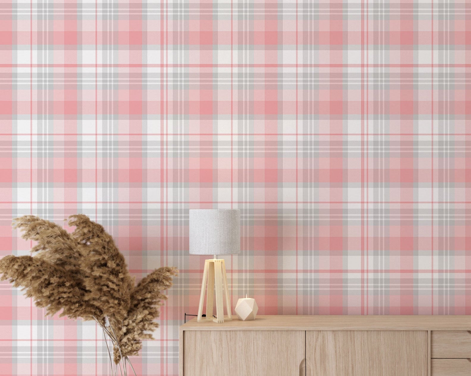 A modern living space featuring a soft cherry blossom pink, white, and grey plaid wallpaper accent wall. In the center, a light oak sideboard cabinet with wooden legs holds a small wooden tripod lamp, a geometric candle, and a round white ceramic vase with a single sprig of dried pampas grass. To the left, a woven basket sits on the light wood floor, filled with tall, fluffy pampas grass stalks and a draped blanket. To the right, a contemporary grey accent chair is styled with a dark throw blanket.