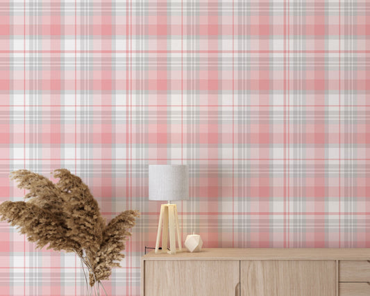A modern living space featuring a soft cherry blossom pink, white, and grey plaid wallpaper accent wall. In the center, a light oak sideboard cabinet with wooden legs holds a small wooden tripod lamp, a geometric candle, and a round white ceramic vase with a single sprig of dried pampas grass. To the left, a woven basket sits on the light wood floor, filled with tall, fluffy pampas grass stalks and a draped blanket. To the right, a contemporary grey accent chair is styled with a dark throw blanket.