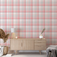 A modern living space featuring a soft cherry blossom pink, white, and grey plaid wallpaper accent wall. In the center, a light oak sideboard cabinet with wooden legs holds a small wooden tripod lamp, a geometric candle, and a round white ceramic vase with a single sprig of dried pampas grass. To the left, a woven basket sits on the light wood floor, filled with tall, fluffy pampas grass stalks and a draped blanket. To the right, a contemporary grey accent chair is styled with a dark throw blanket.