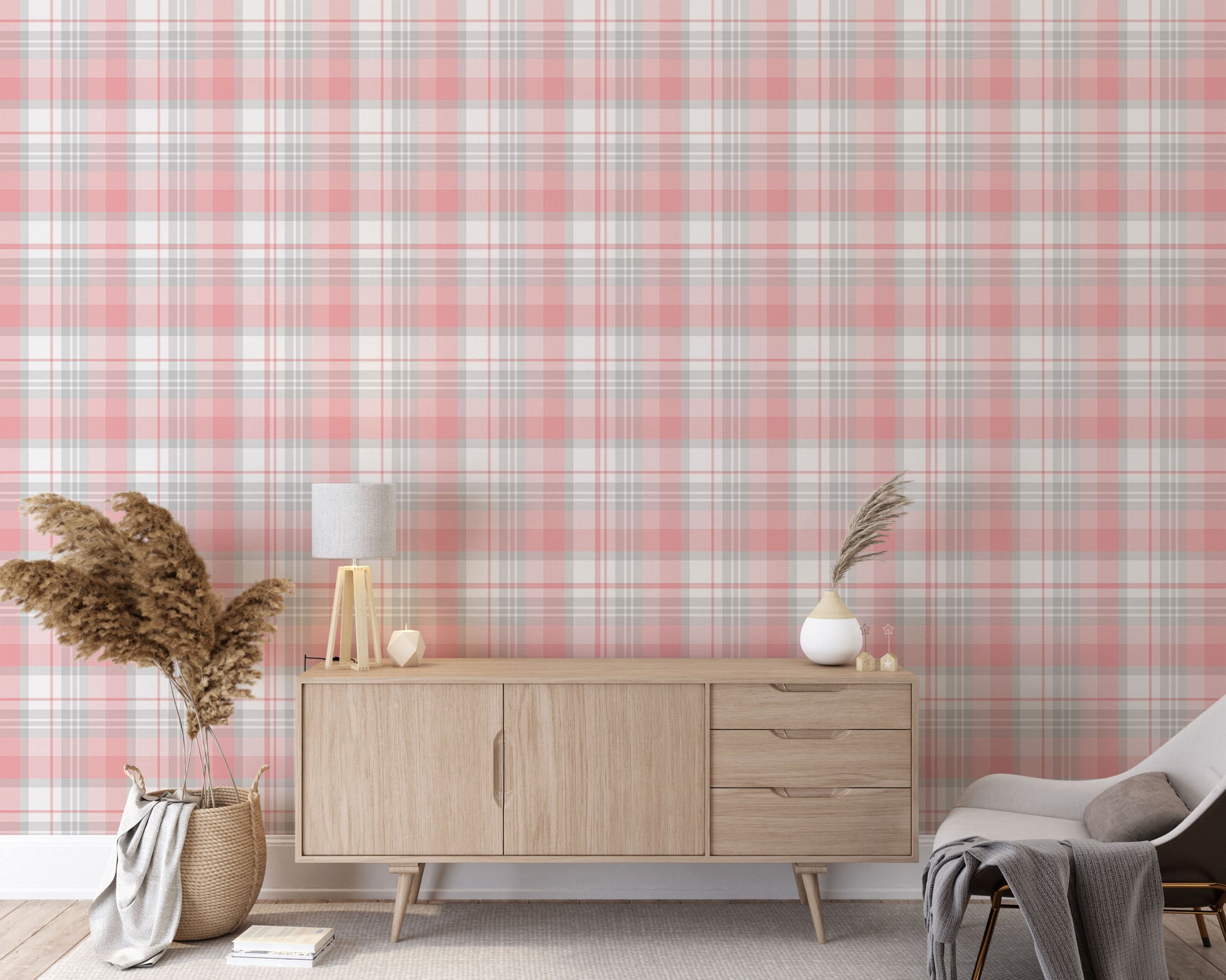 A modern living space featuring a soft cherry blossom pink, white, and grey plaid wallpaper accent wall. In the center, a light oak sideboard cabinet with wooden legs holds a small wooden tripod lamp, a geometric candle, and a round white ceramic vase with a single sprig of dried pampas grass. To the left, a woven basket sits on the light wood floor, filled with tall, fluffy pampas grass stalks and a draped blanket. To the right, a contemporary grey accent chair is styled with a dark throw blanket.