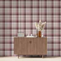 A modern living space featuring a deep burgundy, mauve, and white plaid wallpaper accent wall. Centered against the wall is a mid-century modern wooden sideboard cabinet with vertical fluted doors and tapered legs. On top of the cabinet rests a vintage-style portable radio on the left, a small stack of white books with a geometric sphere on them in the center, and a collection of amber and white glass bottles to the right, filled with dried pampas grass and reed diffusers. 