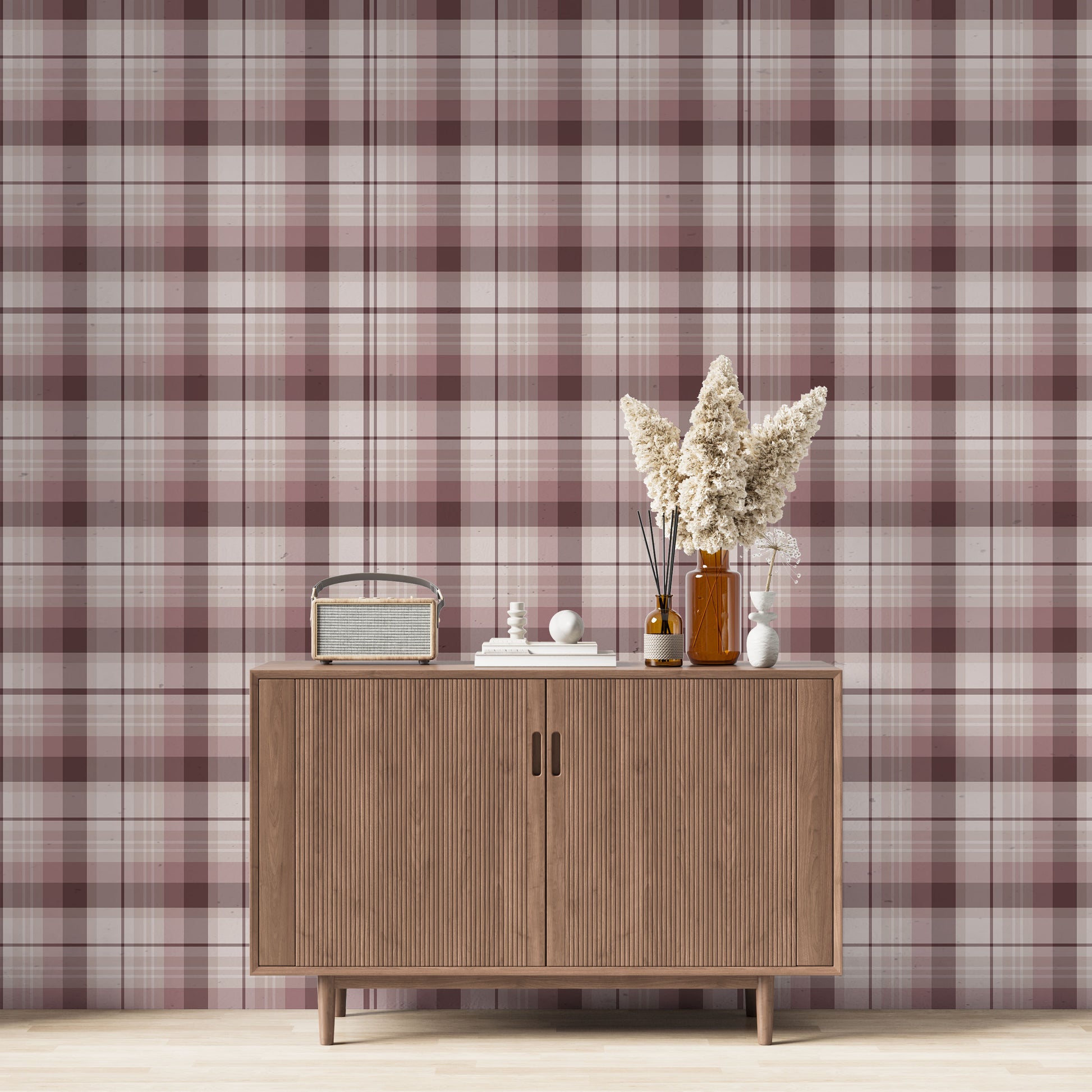 A modern living space featuring a deep burgundy, mauve, and white plaid wallpaper accent wall. Centered against the wall is a mid-century modern wooden sideboard cabinet with vertical fluted doors and tapered legs. On top of the cabinet rests a vintage-style portable radio on the left, a small stack of white books with a geometric sphere on them in the center, and a collection of amber and white glass bottles to the right, filled with dried pampas grass and reed diffusers. 