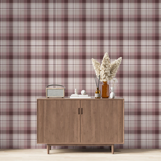 A modern living space featuring a deep burgundy, mauve, and white plaid wallpaper accent wall. Centered against the wall is a mid-century modern wooden sideboard cabinet with vertical fluted doors and tapered legs. On top of the cabinet rests a vintage-style portable radio on the left, a small stack of white books with a geometric sphere on them in the center, and a collection of amber and white glass bottles to the right, filled with dried pampas grass and reed diffusers. 