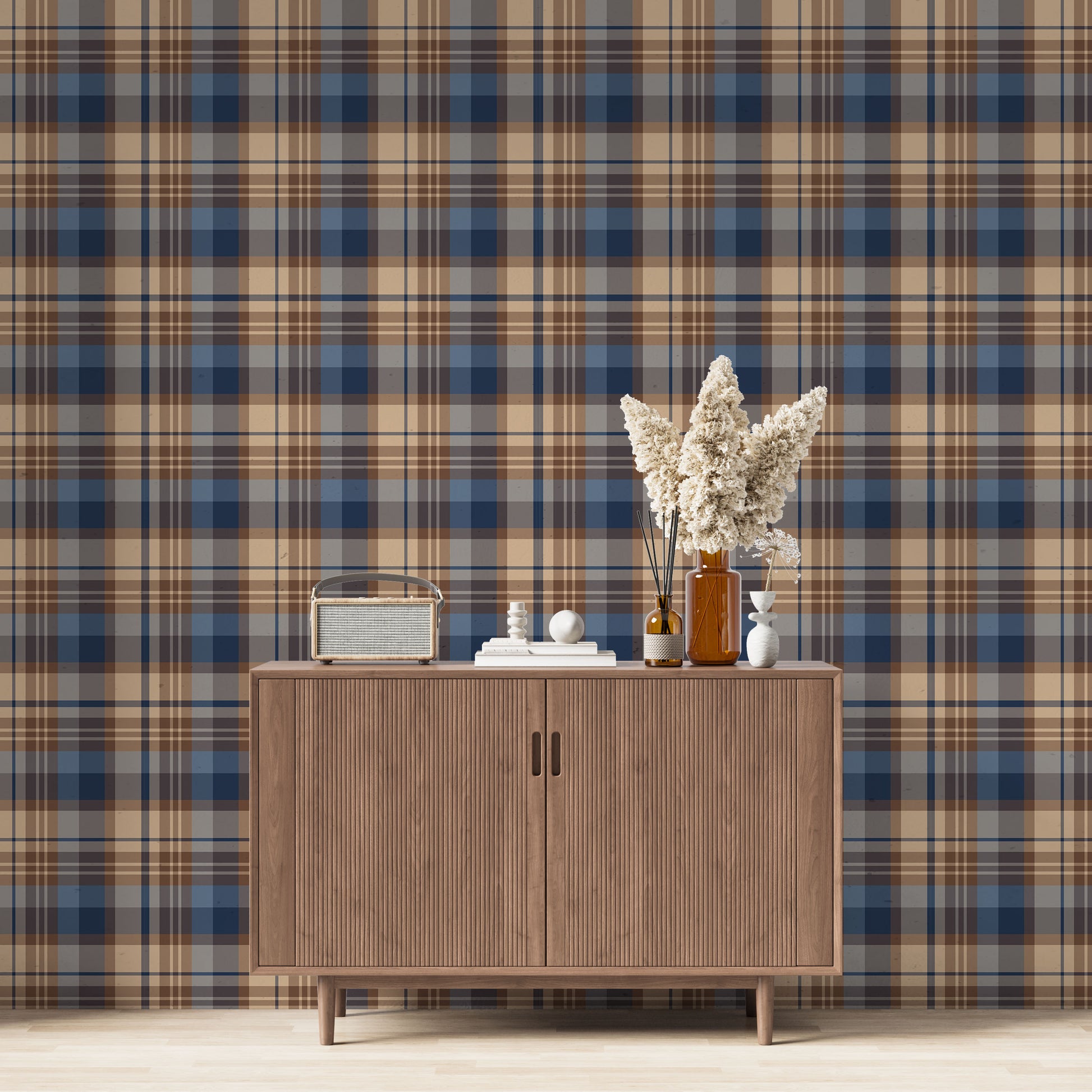 A modern living space featuring a bold navy blue, slate blue, and warm tan plaid wallpaper accent wall. Centered against the wall is a mid-century modern wooden sideboard cabinet with vertical fluted doors and tapered legs. On top of the cabinet rests a vintage-style portable radio on the left, a small stack of white books with a geometric sphere on them in the center, and a collection of amber and white glass bottles to the right, filled with dried pampas grass and reed diffusers.