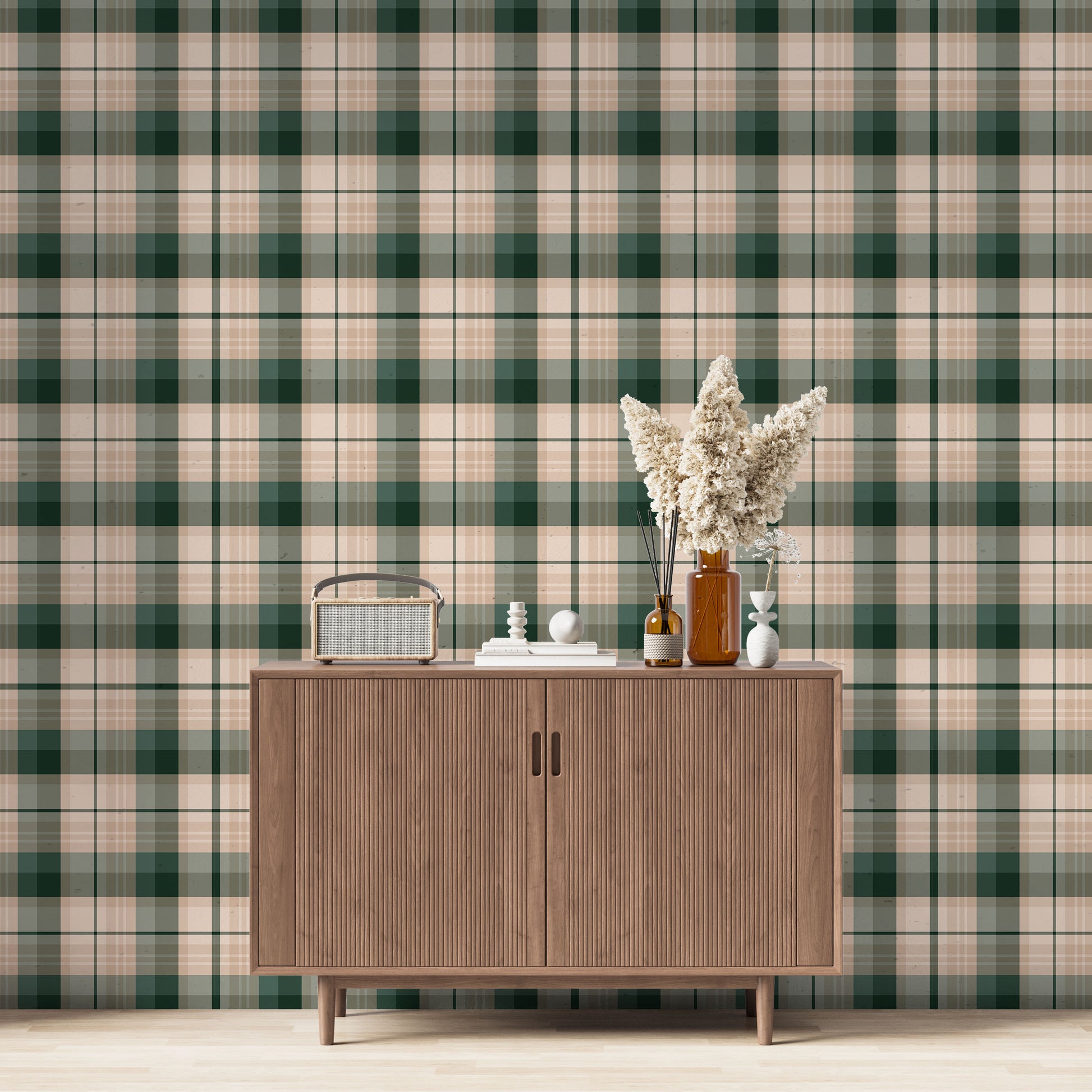 A modern living space featuring a deep forest green, sage green, and peach-tan plaid wallpaper accent wall. Centered against the wall is a mid-century modern wooden sideboard cabinet with vertical fluted doors and tapered legs. On top of the cabinet rests a vintage-style portable radio on the left, a small stack of white books with a geometric sphere on them in the center, and a collection of amber and white glass bottles to the right, filled with dried pampas grass and reed diffusers.