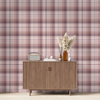 A modern living space featuring a soft rose pink, dusty mauve, and plum plaid wallpaper accent wall. Centered against the wall is a mid-century modern wooden sideboard cabinet with vertical fluted doors and tapered legs. On top of the cabinet rests a vintage-style portable radio on the left, a small stack of white books with a geometric sphere on them in the center, and a collection of amber and white glass bottles to the right, filled with dried pampas grass and reed diffusers.