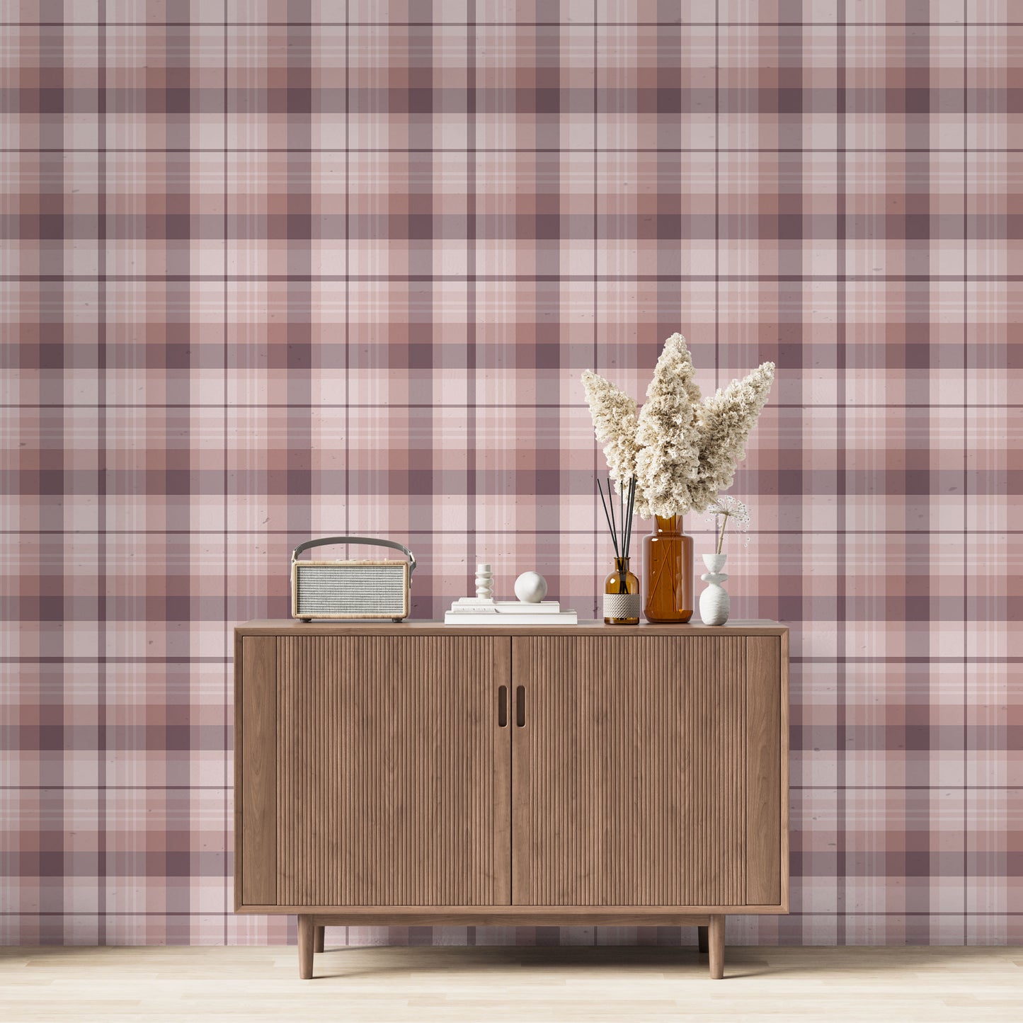 A modern living space featuring a soft rose pink, dusty mauve, and plum plaid wallpaper accent wall. Centered against the wall is a mid-century modern wooden sideboard cabinet with vertical fluted doors and tapered legs. On top of the cabinet rests a vintage-style portable radio on the left, a small stack of white books with a geometric sphere on them in the center, and a collection of amber and white glass bottles to the right, filled with dried pampas grass and reed diffusers.