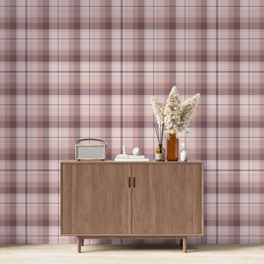 A modern living space featuring a soft rose pink, dusty mauve, and plum plaid wallpaper accent wall. Centered against the wall is a mid-century modern wooden sideboard cabinet with vertical fluted doors and tapered legs. On top of the cabinet rests a vintage-style portable radio on the left, a small stack of white books with a geometric sphere on them in the center, and a collection of amber and white glass bottles to the right, filled with dried pampas grass and reed diffusers.