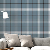 A modern living room features a serene blue-steel grey and white plaid wallpaper.
The detailed design shows intersecting white and dark lines creating grids on a blue base.
In front, a light grey fabric sofa with matching cushions sits centered in the room.
The bright space is complete with a wooden armchair, coffee tables, and a floor lamp.