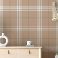 A stylish living area features a warm taupe brown and white plaid wallpaper.
The detailed design shows intersecting white and brown lines creating grids on a taupe base.
In front, a cream-colored wooden sideboard with woven rattan doors is centered nicely.
The serene setup is accented with a white desk lamp, pottery, and a leafy green plant.
