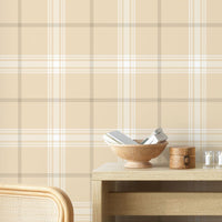 A stylish living area features a soft pale almond and off-white plaid wallpaper.
The detailed design shows intersecting light brown lines forming grids over a sandy base.
In front, a cream-colored wooden sideboard with woven rattan doors is centered nicely.
The serene setup is accented with a white desk lamp, pottery, and a leafy green plant.