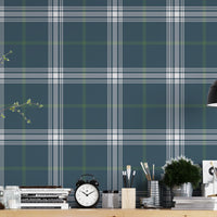 A creative workspace features a deep stormy blue and white plaid wallpaper.
The detailed pattern shows intersecting white and green lines forming grids over a dark base.
In front, a wooden trestle desk holds books, an alarm clock, and art supplies.
The organized setup is completed with a industrial swivel stool and a black pendant light.