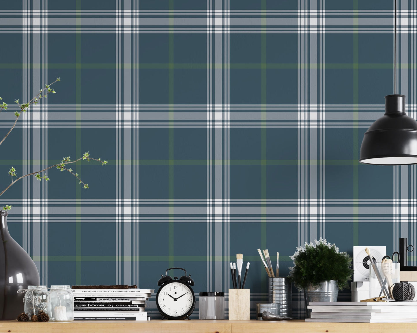 A creative workspace features a deep stormy blue and white plaid wallpaper.
The detailed pattern shows intersecting white and green lines forming grids over a dark base.
In front, a wooden trestle desk holds books, an alarm clock, and art supplies.
The organized setup is completed with a industrial swivel stool and a black pendant light.