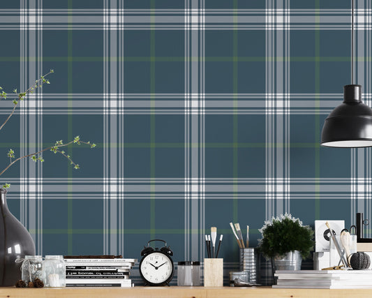 A creative workspace features a deep stormy blue and white plaid wallpaper.
The detailed pattern shows intersecting white and green lines forming grids over a dark base.
In front, a wooden trestle desk holds books, an alarm clock, and art supplies.
The organized setup is completed with a industrial swivel stool and a black pendant light.