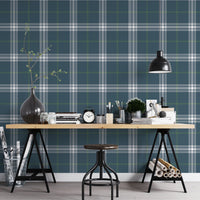 A creative workspace features a deep stormy blue and white plaid wallpaper.
The detailed pattern shows intersecting white and green lines forming grids over a dark base.
In front, a wooden trestle desk holds books, an alarm clock, and art supplies.
The organized setup is completed with a industrial swivel stool and a black pendant light.