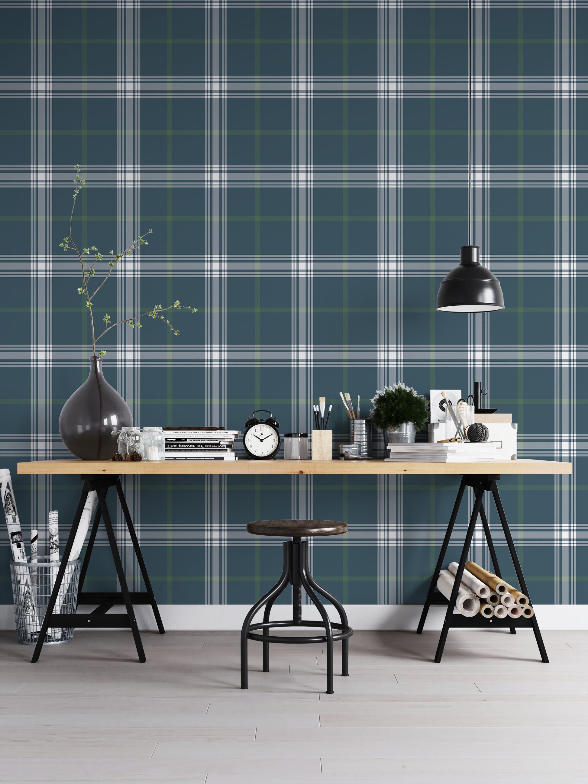 A creative workspace features a deep stormy blue and white plaid wallpaper.
The detailed pattern shows intersecting white and green lines forming grids over a dark base.
In front, a wooden trestle desk holds books, an alarm clock, and art supplies.
The organized setup is completed with a industrial swivel stool and a black pendant light.