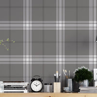 A creative workspace features a dark midnight grey and white plaid wallpaper.
The detailed pattern shows intersecting white and light grey lines forming grids over a dark base.
In front, a wooden trestle desk holds books, an alarm clock, and art supplies.
The organized setup is completed with an industrial swivel stool and a black pendant light.