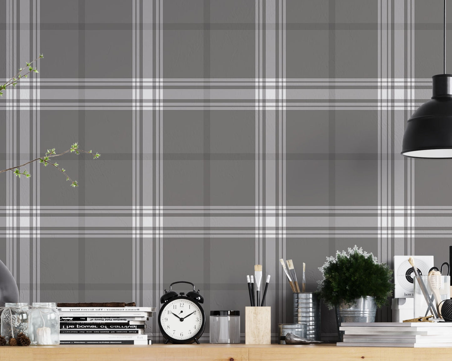 A creative workspace features a dark midnight grey and white plaid wallpaper.
The detailed pattern shows intersecting white and light grey lines forming grids over a dark base.
In front, a wooden trestle desk holds books, an alarm clock, and art supplies.
The organized setup is completed with an industrial swivel stool and a black pendant light.
