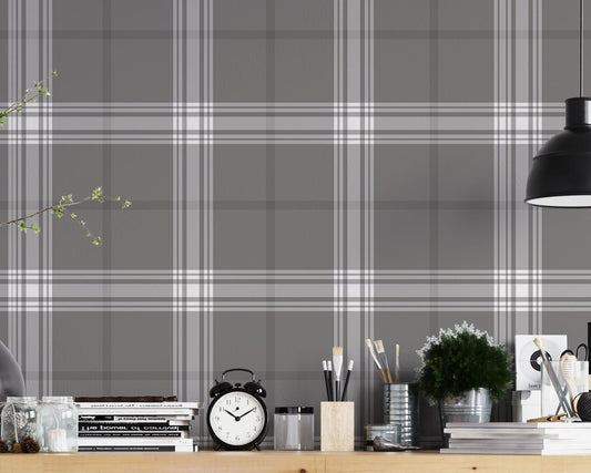 A creative workspace features a dark midnight grey and white plaid wallpaper.
The detailed pattern shows intersecting white and light grey lines forming grids over a dark base.
In front, a wooden trestle desk holds books, an alarm clock, and art supplies.
The organized setup is completed with an industrial swivel stool and a black pendant light.