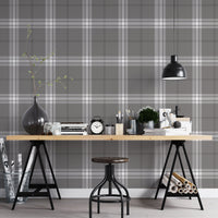 A creative workspace features a dark midnight grey and white plaid wallpaper.
The detailed pattern shows intersecting white and light grey lines forming grids over a dark base.
In front, a wooden trestle desk holds books, an alarm clock, and art supplies.
The organized setup is completed with an industrial swivel stool and a black pendant light.