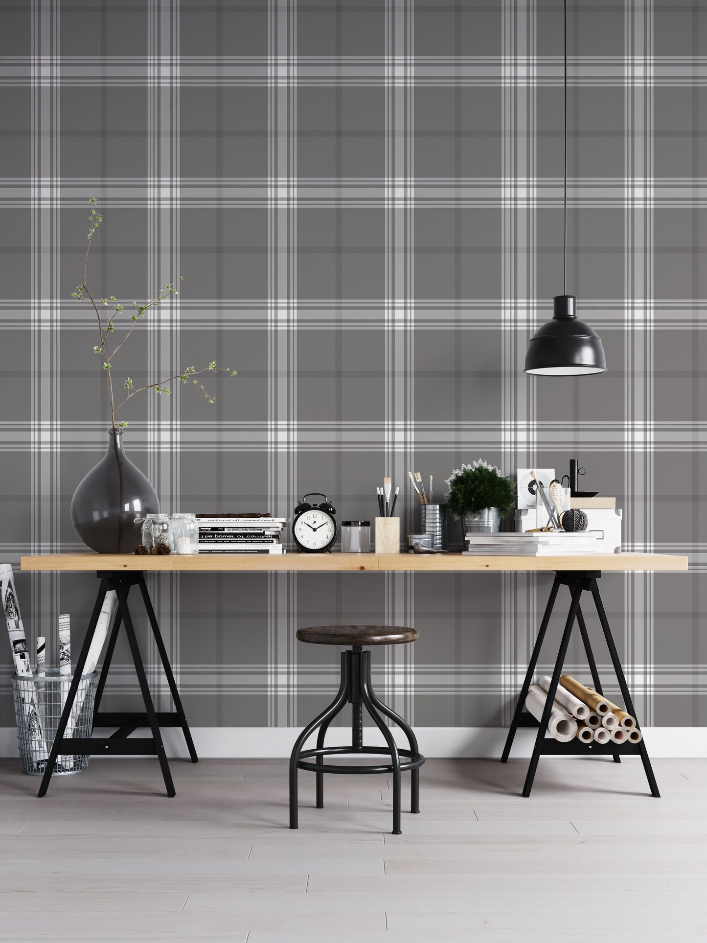 A creative workspace features a dark midnight grey and white plaid wallpaper.
The detailed pattern shows intersecting white and light grey lines forming grids over a dark base.
In front, a wooden trestle desk holds books, an alarm clock, and art supplies.
The organized setup is completed with an industrial swivel stool and a black pendant light.