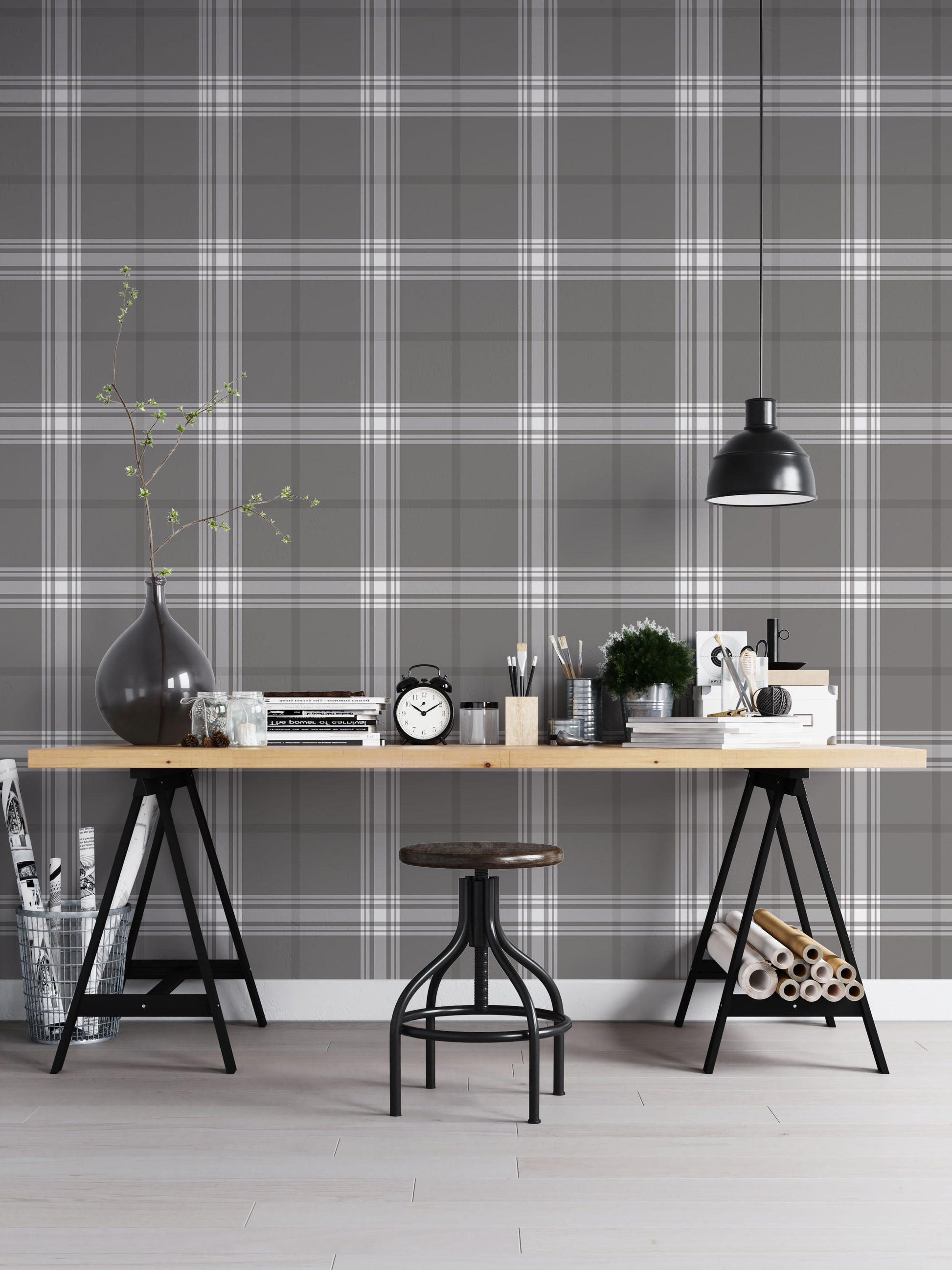 A creative workspace features a dark midnight grey and white plaid wallpaper.
The detailed pattern shows intersecting white and light grey lines forming grids over a dark base.
In front, a wooden trestle desk holds books, an alarm clock, and art supplies.
The organized setup is completed with an industrial swivel stool and a black pendant light.