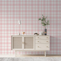 A charming living area features a soft baby pink and white plaid wallpaper.
The detailed design shows intersecting light pink lines forming grids over a light base.
In front, a cream-colored wooden sideboard with woven rattan doors is centered nicely.
The serene setup is accented with a white desk lamp, pottery, and a leafy green plant.