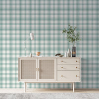 A serene living area features a soft baby green and white plaid wallpaper.
The detailed design shows intersecting light green lines forming grids over a light base.
In front, a cream-colored wooden sideboard with woven rattan doors is centered nicely.
The relaxing setup is accented with a white desk lamp, pottery, and a leafy green plant.