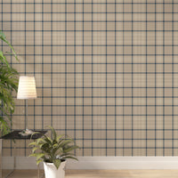 A modern living space featuring a warm soft caramel brown, beige, and cream plaid wallpaper accent wall. In the center, a light oak sideboard cabinet with wooden legs holds a small wooden tripod lamp, a geometric candle, and a round white ceramic vase with a single sprig of dried pampas grass. To the left, a woven basket sits on the light wood floor, filled with tall, fluffy pampas grass stalks and a draped blanket.
