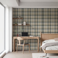 A closeup of a modern bedroom and study area featuring a prominent stone-beige plaid wallpaper. On the left, a light oak wooden desk holds a closed laptop, a small stack of books, and a sleek, matte black metal desk lamp. To the right, a matching light oak wooden headboard and bedframe are visible, with a crisp white duvet pillow and a soft beige accent pillow resting on top.