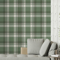 A modern living space featuring a prominent olive green and white plaid wallpaper accent wall. In front of the wall sits a sleek grey chaise lounge with matching plush pillows, accompanied by a small, round metal side table. To the left, a large window is dressed with sheer white curtains and long, sage green drapes over herringbone wood flooring. To the right, a textured white feature wall provides contrast, with a large potted green plant in a dark ceramic planter standing in the corner.